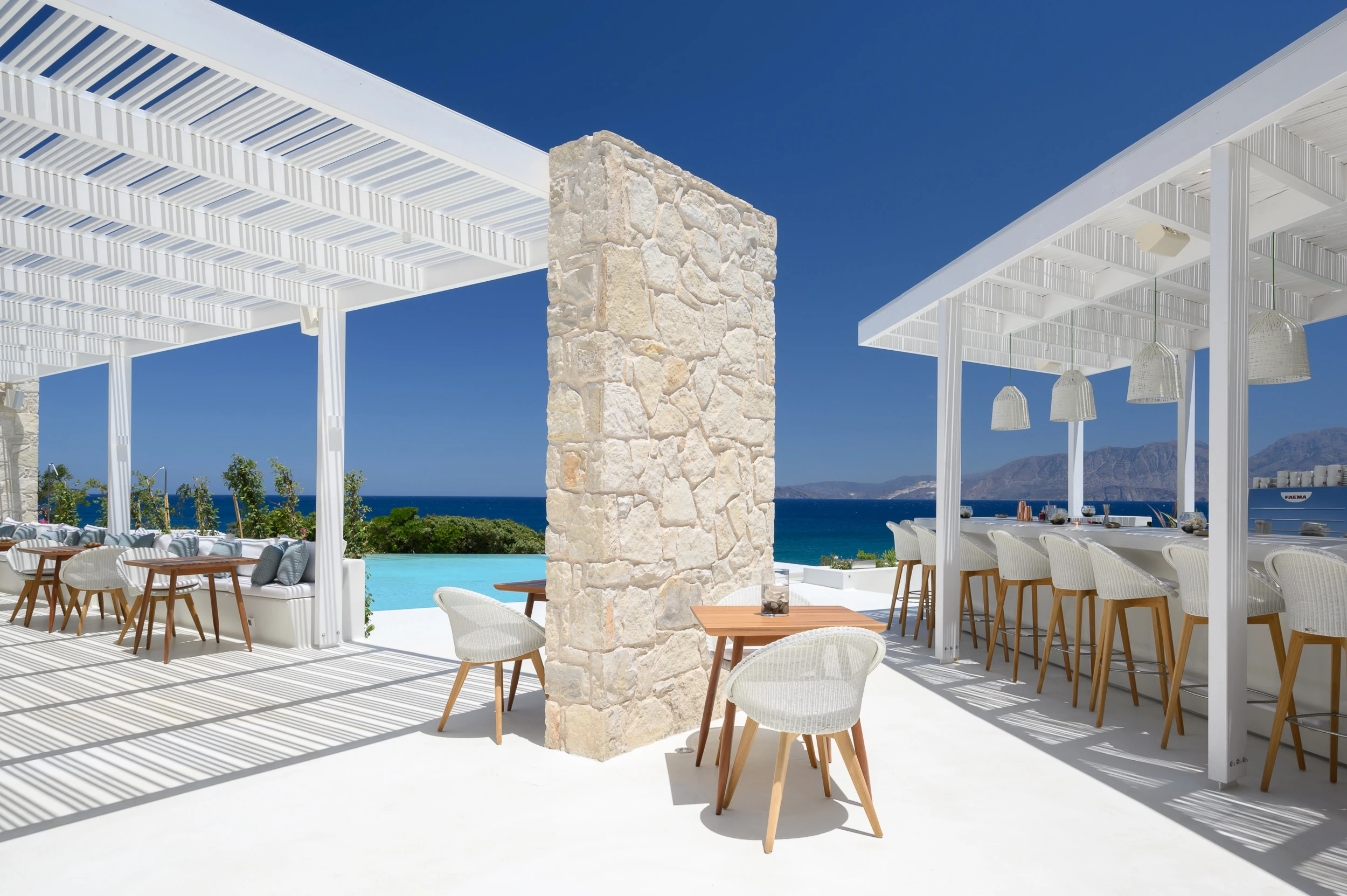 The Island Concept — Πέργκολα και υπαίθρια τραπεζαρία Υπαίθρια τραπεζαρία στο The Island Concept με λευκή πέργκολα, πέτρινη κολόνα και θέα στη θάλασσα.